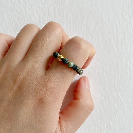 Hand wearing a ring with green beads and gold accents on a white background