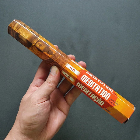 Hand holding a wooden meditation instrument with 'Meditation' text on an orange label against a gray background
