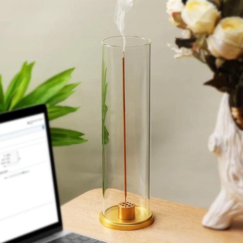 Incense stick in a glass container on a wooden surface with a laptop and flowers in the background.