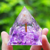 Pyramid-shaped crystal with purple and white stones held by a hand against a green blurred background