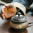 Ceramic incense burner with smoke on a wooden surface, blurred hands in the background.