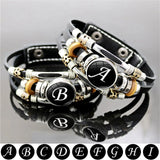 Black leather bracelet with silver and gold charms featuring initials on a gray background.