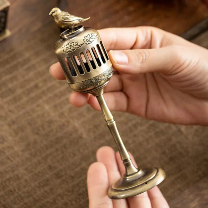 Hand holding a small brass incense burner with a bird design against a blurred background