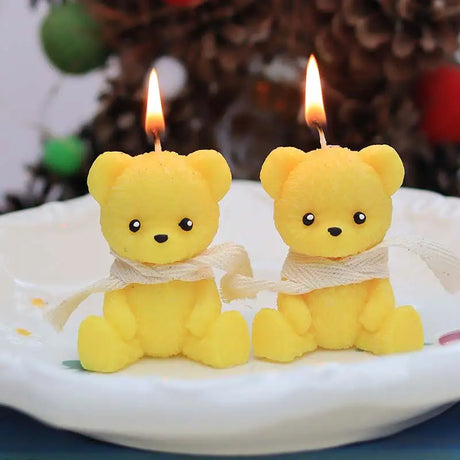 Two yellow bear-shaped candles with lit wicks on a white plate against a blurred background.