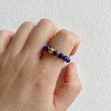 Hand wearing a ring with blue and gold beads on a plain background