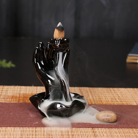 Incense burner shaped like hands holding incense on a mat with a dark background