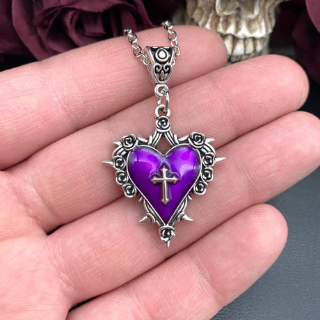 Purple heart-shaped pendant with a cross on a silver chain held in a hand.