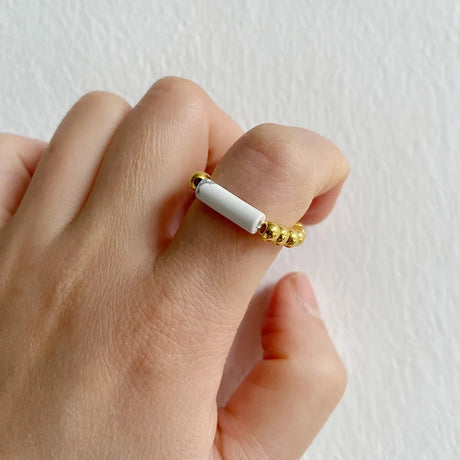 Hand wearing a ring with a white cylindrical center and gold beads on a plain background